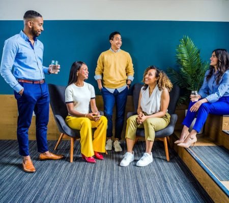 A diverse group of five professionals engaging in a casual indoor meeting, smiling and talking.
